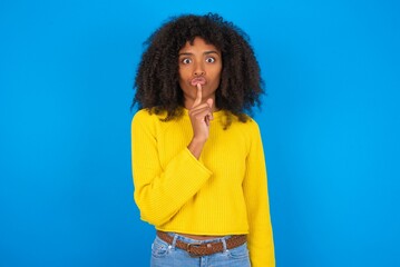 Surprised young woman with afro hairstyle wearing yellow sweater over blue wall makes silence gesture, keeps finger over lips and looks mysterious at camera