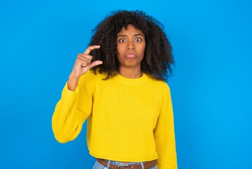 young woman with afro hairstyle wearing yellow sweater over blue wall purses lip and gestures with hand, shows something very little.