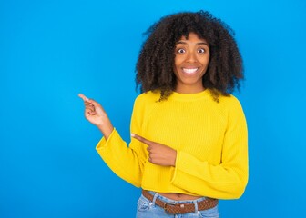 young woman with afro hairstyle wearing yellow sweater over blue wall points at copy space indicates for advertising gives right direction
