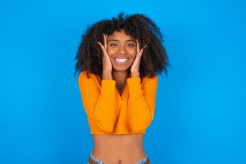 Happy young woman with afro hairstyle wearing orange crop top over blue wall touches both cheeks gently, has tender smile, shows white teeth, gazes positively straightly at camera,