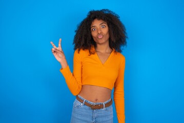 young beautiful brunette woman wearing colourful dress over white wall makes peace gesture keeps...