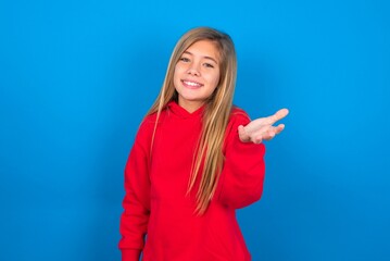 caucasian teen girl wearing red sweater over blue studio background smiling friendly offering something with open hand or handshake as greeting and welcoming. Successful business.
