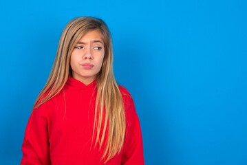 caucasian teen girl wearing red sweater over blue studio background , looks pensively aside, plans actions after university, imagines what to do Thinks over about new project.