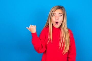Shocked caucasian teen girl wearing red sweater over blue studio background points with thumb away, indicates something. Check this out. Advertisement concept.