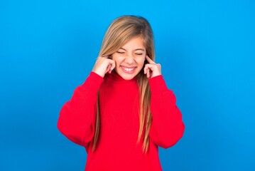 Stop making this annoying sound! Unhappy stressed out caucasian teen girl wearing red sweater over blue studio background making worry face, plugging ears with fingers, irritated with loud noise.