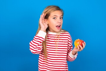 Fototapeta na wymiar Oh my God! Funny astonished caucasian teen girl wearing striped shirt over blue studio background opening mouth widely looking aside, with hand near ear trying to listen to gossips.
