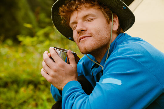 Young man drinking tea while sitting at camping tent in forest - Powered by Adobe
