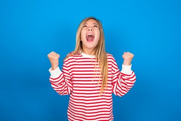 caucasian teen girl wearing striped shirt over blue studio background looks with excitement up, keeps hands raised, notices something unexpected.