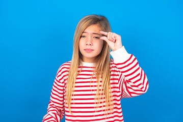 Upset caucasian teen girl wearing striped shirt over blue studio background shapes little gesture with hand demonstrates something very tiny small size. Not very much