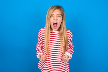 Joyful excited lucky caucasian teen girl wearing striped shirt over blue studio background cheering, celebrating success, screaming yes with clenched fists