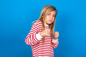 Portrait of attractive caucasian teen girl wearing striped shirt over blue studio background holding hands in front of him in boxing position going to fight.