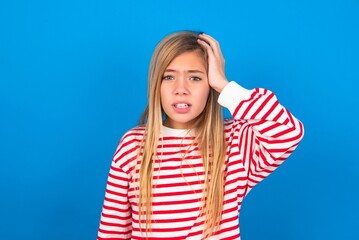 Portrait of confused caucasian teen girl wearing striped shirt over blue studio background holding hand on hair and frowning, panicking, losing memory. Worried and anxious can not remember anything.