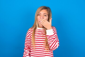 Sleepy caucasian teen girl wearing striped shirt over blue studio background yawning with messy hair, feeling tired after sleepless night, yawning, covering mouth with palm.