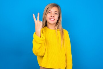 caucasian teen girl wearing yellow sweater over blue studio background smiling and looking friendly, showing number two or second with hand forward, counting down
