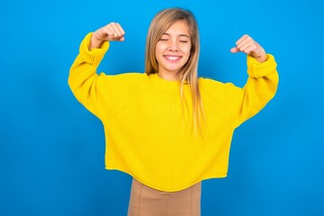Strong powerful caucasian teen girl wearing yellow sweater over blue studio background toothy...
