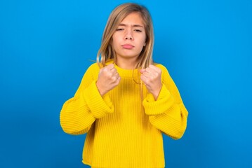 Displeased annoyed caucasian teen girl wearing yellow sweater over blue studio clenches fists, gestures pissed, ready to revenge, looks with aggression at camera stands full of hate, being pressured