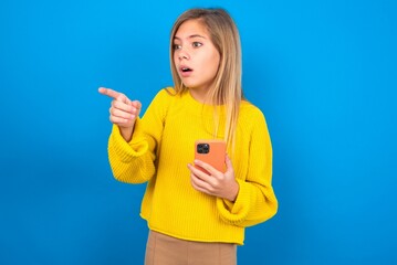 Stunned caucasian teen girl wearing yellow sweater over blue studio background points sideways right copy space, recommends product, sees astonishing thing