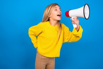 Funny caucasian teen girl wearing yellow sweater over blue studio background People sincere emotions lifestyle concept. Mock up copy space. Screaming in megaphone.