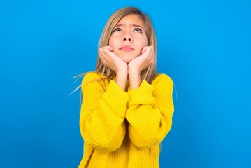 Portrait of sad caucasian teen girl wearing yellow sweater over blue studio background hands face...
