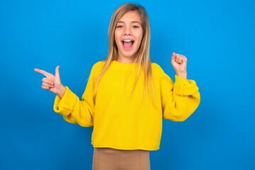 caucasian teen girl wearing yellow sweater over blue studio background points at empty space holding fist up, winner gesture.