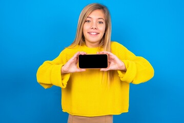 Cheerful cheery content caucasian teen girl wearing yellow sweater over blue studio background holding in hands device hobby smm post blog