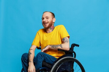 A man in a wheelchair smile looks at the camera in a t-shirt with tattoos on his arms sits on a blue studio background, a full life, a real person
