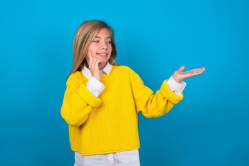 Funny caucasian teen girl wearing yellow sweater over blue studio background holding open palm new product. I wanna buy it!