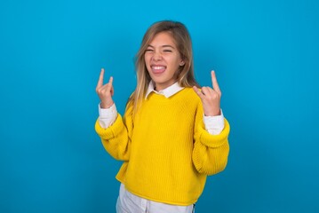 caucasian teen girl wearing yellow sweater over blue studio background making rock hand gesture and...
