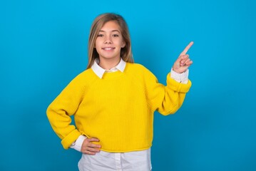 caucasian teen girl wearing yellow sweater over blue studio background looking at camera indicating finger empty space sales