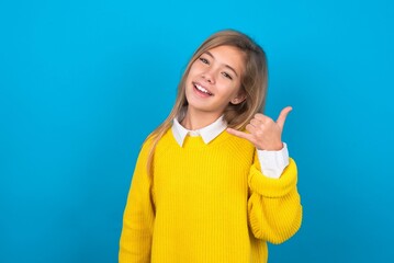 caucasian teen girl wearing yellow sweater over blue studio background smiling doing phone gesture with hand and fingers like talking on the telephone. Communicating concepts.