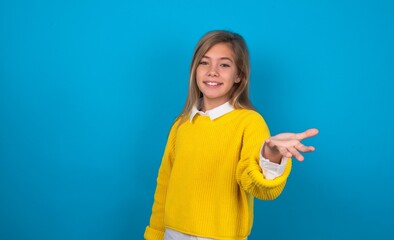 caucasian teen girl wearing yellow sweater over blue studio background smiling cheerful offering palm hand giving assistance and acceptance.