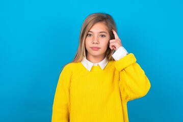caucasian teen girl wearing yellow sweater over blue studio background tries to memorize something,...