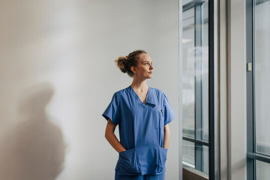 Thoughtful Young Nurse Standing With Hands In Pockets Against Wall At Hospital