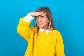 caucasian teen girl wearing yellow sweater over blue studio background very happy and smiling looking far away with hand over head. Searching concept.