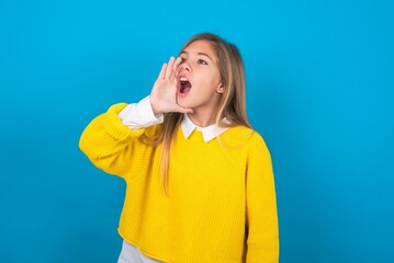 caucasian teen girl wearing yellow sweater over blue studio background shouting and screaming loud to side with hand on mouth. Communication concept.