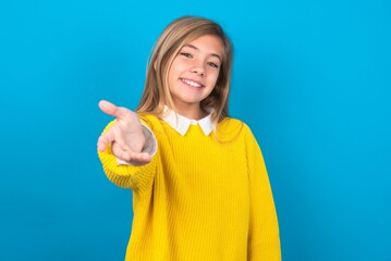 caucasian teen girl wearing yellow sweater over blue studio background smiling friendly offering handshake as greeting and welcoming. Successful business.