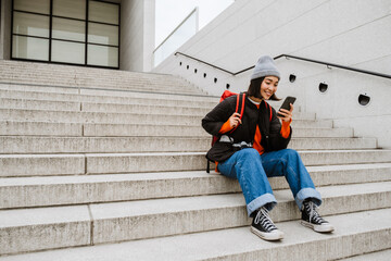 Positive asian woman using mobile phone while sitting on stairs at city street - obrazy, fototapety, plakaty