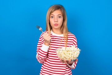 hungry beautiful caucasian teen girl wearing striped T-shirt over blue wall eating popcorn holding in hand fork knife want tasty yummy pizza pie