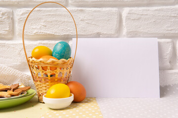 Basket with Easter colored eggs, plate with holiday cookies and white empty paper card