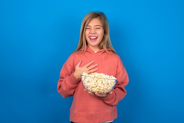 beautiful caucasian teen girl wearing red sweater over blue wall eating popcorn expresses happiness, laughs pleasantly, keeps hands on heart