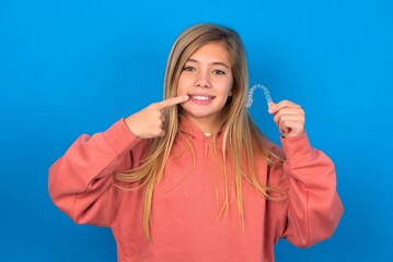 Fototapeta premium beautiful caucasian teen girl wearing pink sweater over blue wall holding an invisible aligner and pointing perfect straight teeth. Dental healthcare and confidence concept.