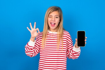 beautiful caucasian teen girl wearing striped t-shirt over blue background holding in hands cell...