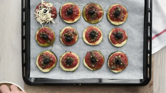 Manos de mujer ponen queso rallado sobre aceitunas alcaparras, rodajas de berenjena con or&eacute;gano, aceite y tomate natural triturado en una bandeja de horno
