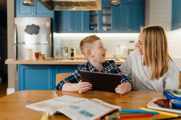 Cheerful mom and son using tablet computer while sitting in kitchen