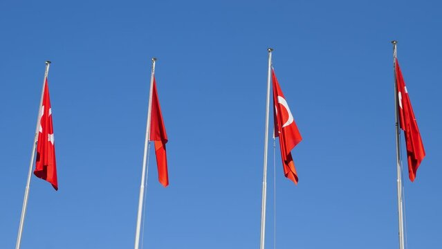 Four Turkish flags hang from flagpoles in small wind weather, low angle shot against blue sky. Red fabric slightly move, but wind is too weak to show whole sign