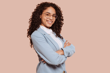Young smiling successful mixed race woman entrepreneur or an office worker stands with crossed arms at studio isolated over beige background.