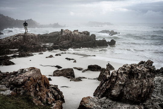 A man standing on a rock in misty overcast conditions along the Walker Bay coastline. Hermanus, Whale Coast, Overberg, Western Cape, South Africa.