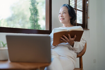 Portrait of an elderly Asian woman in a modern pose holding a memory notebook and operating a computer.