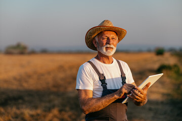 An elderly man stands in a field being plowed, holding a digital tablet that helps him monitor farm machinery. In the background is the sunset.