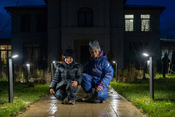 Obraz premium Two happy sisters pose on path in front of house illuminated by row of modern street lamps on rainy evening backyard